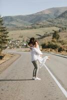 ein stilvoll Bräutigam im ein Weiß Hemd und ein süß Brünette Braut im ein Weiß Kleid sind kreisen auf ein Asphalt Straße gegen das Hintergrund von ein Wald und Berge. Hochzeit Porträt von Jungvermählten. foto