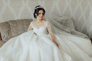 ein Brünette Braut im ein Weiß voluminös Kleid mit lange Spitze Ärmel und ein schick Krone posiert während Sitzung auf ein grau Sofa im ihr Zimmer. großartig bilden. öffnen Schultern. schön Hände. foto