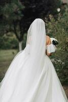 Hochzeit Foto. das Braut im ein voluminös Weiß Kleid und ein lange Schleier Spaziergänge im das Garten entlang ein Stein Weg, halten ein Strauß von Weiß Rosen, ihr Schultern sind gedreht zu das Kamera. foto