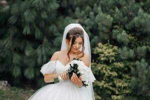 Brünette Braut im ein voluminös Weiß Kleid mit öffnen Schultern und ein lange Schleier, lächelnd, posieren mit ein Strauß von Weiß Rosen. Porträt von das Braut. schön bilden und Haar. foto