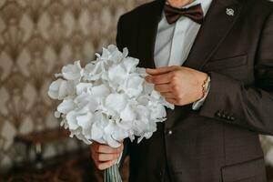 das Bräutigam im ein braun Jacke und Weiß Hemd mit ein Strauß von Blumen im ein Zimmer mit ein wunderbar Innere. das Bräutigam sieht aus beim das Strauß foto