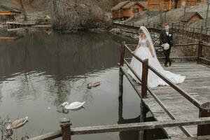 ein stilvoll Braut im ein üppig Kleid und modisch Frisur steht auf ein Seebrücke im ein Park in der Nähe von hölzern Häuser, das Bräutigam hinter ihr. Schwäne schwimmen im das See foto