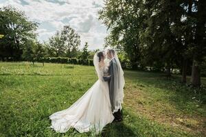 ein lockiges Haar Brünette Braut im ein Weiß Kleid und ein verschleiert Bräutigam Umarmung und Kuss. Porträt von das Braut und Bräutigam. schön bilden und Haar. Hochzeit im Natur foto