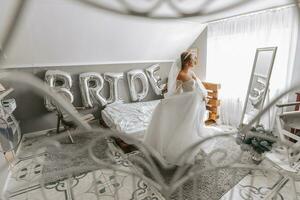 schön lockig braunhaarig Braut im ein Weiß Kleid posiert zum ein Fotograf, kreisen das Zimmer im ein schön Kleid mit Ärmel. Hochzeit Fotografie, Nahansicht Porträt, schick Frisur. foto