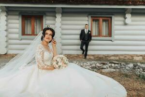 ein Brünette Braut mit ein Krone auf ihr Kopf im ein Weiß voluminös Kleid ist posieren, halten ein Strauß im ihr Hände, das Bräutigam ist Stehen hinter ihr gelehnt auf ein Weiß Hütte. Hochzeit Schießen foto