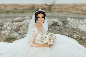 Porträt von das Braut im Natur. ein Brünette Braut mit ein Krone auf ihr Kopf, im ein Weiß voluminös Kleid sitzt, Posen, halten ein Strauß im ihr Hände. schön Haar und bilden. Hochzeit Schießen foto