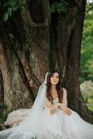 schön Braut mit lange lockig Haar im ein schick Kleid, lächelnd, suchen in das Linse unter ein groß Baum. Porträt von das Braut. Frühling Hochzeit. natürlich bilden foto