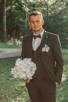 jung elegant Bräutigam im das Wald mit ein Strauß von Weiß Blumen im ein braun passen und ein Bogen Krawatte foto