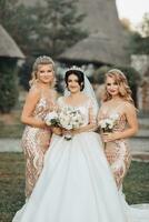 ein Brünette Braut im ein Weiß elegant Kleid mit ein Krone und ihr blond freunde im Gold Kleider Pose mit Blumensträuße. Hochzeit Porträt im Natur, Hochzeit Foto im ein Licht Ton.