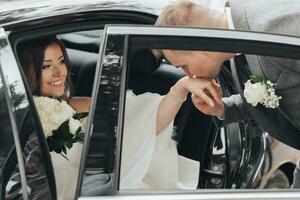 Hochzeit Foto. das Braut sitzt im ein schwarz Auto mit ein lächeln, das Bräutigam Küsse das Braut Hand. Aussicht durch das Auto Fenster. schön bilden und Haar. ein luxuriös Weiß Kleid foto