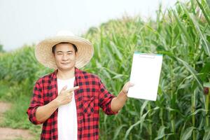 gut aussehend asiatisch Mann Farmer trägt Hut, rot Plaid Shirt, halt und Punkt zu leer Papier Zwischenablage, steht beim Mais Garten. Konzept. Landwirtschaft Beruf, Umfrage und Forschung foto