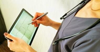 Schuss von das weiblich Arzt im Uniform gegen das Weiß Mauer studieren Kardiogramm auf das Tablette. Gesundheitswesen foto