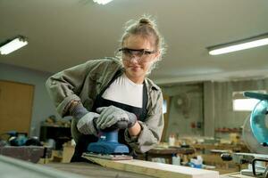 weiblich Zimmermann tragen schützend Sicherheit Brille und mit elektrisch Arbeit auf ein Holz. Künstler oder Möbel Designer Arbeiten auf ein Produkt Idee im ein Werkstatt. foto