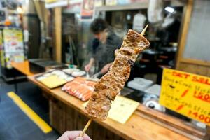 Nahansicht Hand von Tourist halten und Essen Grill kobe Rindfleisch Holz Stock auf verschwommen Grill kobe Rindfleisch Geschäft beim kuromon Markt, Osaka, Japan. foto