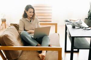 froh tausendjährig Frau Tippen auf ein Laptop, Teilnahme ein virtuell treffen, sitzend auf ein Sofa. heiter tausendjährig weiblich Tippen auf Laptop, hat Treffen mit Gerät, sitzen auf Sofa foto