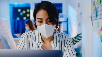 Fröhliche Geschäftsfrau in Asien, die eine medizinische Gesichtsmaske für soziale Distanzierung in einer neuen normalen Situation zur Virenprävention trägt, während sie in der Büronacht den Laptop bei der Arbeit verwendet. Leben und Arbeiten nach dem Coronavirus.