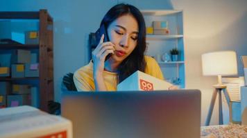 Junge Asiatin ruft Smartphone-Gespräch mit dem Kunden an, um die Bestellung auf Lager auf dem Laptop im Home Office in der Nacht zu überprüfen. Kleinunternehmen, Online-Marktlieferung, Lifestyle-Freelance-Konzept. foto