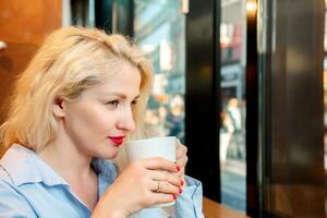 Porträt von ein jung Frau Trinken Kaffee oder Tee im ein Cafe. das Mädchen ist gekleidet im ein Blau Shirt, blond. Sitzung seitwärts und suchen aus das Fenster foto