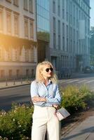 ein jung elegant Frau gekleidet im ein Anzug, Stehen im ein groß Stadt, halten ein Tasche. Gehen Nieder das Straße im das öffnen Luft. ein warm Tag, Sommer. hoch bis zur Hüfte Foto