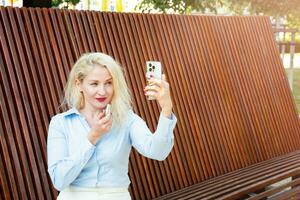 ein nett jung Frau ist Sitzung auf ein Bank im das Park, Gemälde ihr Lippen und reden auf das Telefon, Video Kommunikation. Sommer- Tag. blond foto