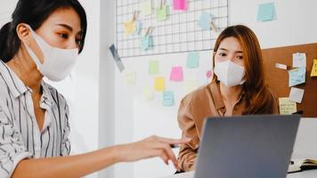 asiatische Geschäftsleute, die Computerpräsentationen verwenden, Brainstorming-Ideen über neue Projektkollegen und tragen eine schützende Gesichtsmaske im neuen normalen Büro. Lebensstil und Arbeit nach dem Coronavirus.