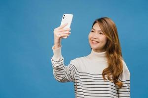 Lächelnde entzückende asiatische Frau, die Selfie-Foto auf Smartphone mit positivem Ausdruck in Freizeitkleidung macht und auf blauem Hintergrund steht. glückliche entzückende frohe frau freut sich über erfolg. foto