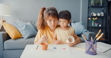 Fröhliche, fröhliche Asien-Familienmutter lehren Kleinkind-Mädchen malen Keramiktopf, der Spaß hat, entspannen Sie sich auf dem Tisch im Wohnzimmer im Haus. Zeit miteinander verbringen, soziale Distanz, Quarantäne zur Coronavirus-Prävention.