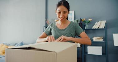 Fröhliche schöne asiatische Dame, die das Karton-Lieferpaket vom Online-Marktplatz im Wohnzimmer im Haus auspackt. Zufriedener Käufer im Internet-Unboxing-Waren-, Online-Shopping- und Lieferkonzept. foto