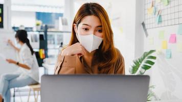 Asiatische Geschäftsfrau trägt Gesichtsmaske für soziale Distanzierung in neuer normaler Situation zur Virusprävention, während sie Kollegen Laptop-Präsentation über den Plan in Videoanrufen während der Arbeit im Büro verwendet. foto