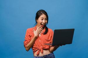 junge asiatische Dame mit Laptop mit positivem Ausdruck, lächelt breit, in Freizeitkleidung gekleidet, fühlt sich glücklich und steht isoliert auf blauem Hintergrund. glückliche entzückende frohe frau freut sich über erfolg. foto