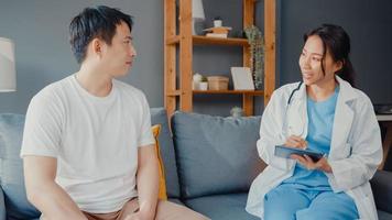 Junge asiatische Ärztin mit digitaler Tablette, die gute Gesundheitstestnachrichten mit glücklichen männlichen Patienten teilt, sitzt auf der Couch im Haus. Krankenversicherung, besuchen Sie den Patienten zu Hause Konzept. foto
