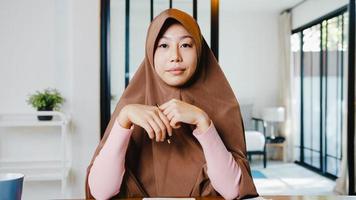 Asiatische muslimische Dame trägt Hijab mit Computer-Laptop Sprechen Sie mit Kollegen über den Plan in einem Videoanruf-Meeting, während Sie von zu Hause aus im Wohnzimmer arbeiten. soziale Distanzierung, Quarantäne wegen Corona-Virus.