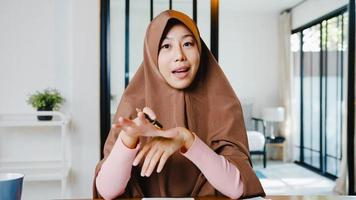 Asiatische muslimische Dame trägt Hijab mit Computer-Laptop Sprechen Sie mit Kollegen über den Plan in einem Videoanruf-Meeting, während Sie von zu Hause aus im Wohnzimmer arbeiten. soziale Distanzierung, Quarantäne wegen Corona-Virus.