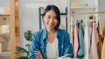 Porträt der jungen asiatischen Modedesignerin, die sich glücklich lächelt, die Arme verschränkt und auf die Kamera schaut, während sie im Bekleidungsgeschäft im Home Office arbeitet. Kleinunternehmer, Online-Markt-Bereitstellungskonzept. foto