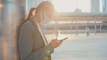 Junge asiatische Geschäftsfrau in modischer Bürokleidung trägt eine medizinische Gesichtsmaske mit dem Telefon, während sie allein im Freien in der Stadt spazieren geht. Geschäft unterwegs, soziale Distanzierung, um die Ausbreitung des Covid-19-Konzepts zu verhindern.