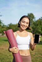 aufgeregt Fitness Mädchen empfiehlt Anwendung zum Sport und trainieren, zeigt an Telefon Bildschirm, Stehen mit Gummi Yoga Matte im Park nach Ausbildung Session foto