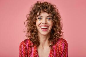 Porträt von glücklich jung Frau mit lockig Haar, lächelt breit, tragen gestreift Kleider mit Muster, isoliert Über Rosa Hintergrund. positiv Emotionen Konzept foto