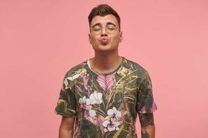 Porträt von süß jung Mann im blühte T-Shirt mit geschlossen Augen, Herstellung Luft Kuss mit seine Lippen, posieren Über Rosa Hintergrund foto