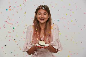 glücklich jung schön lange behaart weiblich zeigen ihr angenehm Emotionen während feiern ihr Geburtstag, lächelnd positiv mit Kuchen im angehoben Hände, posieren Über Weiß Hintergrund foto
