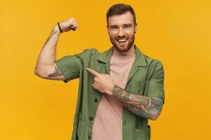 glücklich attraktiv jung Mann mit Bart und tätowieren zeigen und zeigen auf Bizeps Muskeln Über Gelb Hintergrund foto