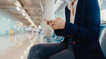 Asiatische Geschäftsfrauenreisende tragen Anzug mit Koffer und verwenden eine Smartphone-Chat-Nachricht in der Bank, um auf den Flug am Flughafen zu warten. Geschäftsreisender Pendler in Covid-Pandemie, Geschäftsreisekonzept. foto