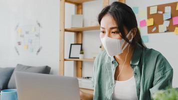 Asiatische Geschäftsfrau, die eine medizinische Gesichtsmaske mit Laptop trägt, spricht mit Kollegen über den Plan im Videoanruf, während sie von zu Hause aus im Wohnzimmer arbeitet. soziale Distanzierung, Quarantäne zur Vorbeugung des Coronavirus.