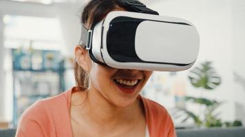 Asiatische Dame mit Headset-Brille der virtuellen Realität, die Hand auf der Couch im Wohnzimmer im Haus gestikuliert. Bleiben Sie zu Hause, Covid-Quarantäne, stellen Sie sich die Realität neu vor, VR-Technologie des zukünftigen Konzepts.