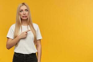 Teenager Mädchen, unzufrieden suchen Frau mit blond lange Haar. tragen Weiß T-Shirt und schwarz Jeans. Menschen und Emotion Konzept. zeigen zu das richtig beim Kopieren Raum mit ein der Ekel Gesichts- Ausdruck foto