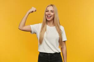 Teenager Mädchen, glücklich suchen Frau mit blond lange Haar. tragen Weiß T-Shirt und schwarz Jeans. zeigt an ihr Muskeln Aufpassen beim das Kamera, isoliert Über Orange Hintergrund foto