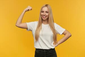 Teenager Mädchen, glücklich suchen Frau mit blond lange Haar. tragen Weiß T-Shirt und schwarz Jeans. zeigt an ihr Muskeln Aufpassen beim das Kamera, isoliert Über Orange Hintergrund foto