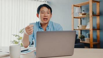 Junger asiatischer Geschäftsmann, der Laptop verwendet, spricht mit Kollegen über den Plan im Videoanruf, während er von zu Hause aus im Wohnzimmer arbeitet. Selbstisolation, soziale Distanzierung, Quarantäne zur Vorbeugung des Coronavirus.