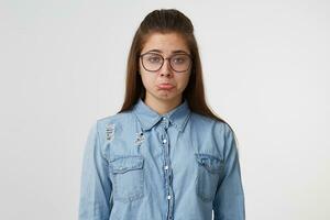 Porträt von ein jung Mädchen tragen Brille mit lange Haar gekleidet im Denim Hemd Schmollmund ihr Lippen, nicht im ein Stimmung, aus von Humor, betrübt, verärgern, isoliert auf Weiß Hintergrund foto