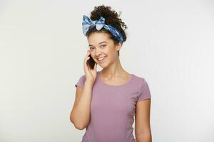 Porträt von nett süß stilvoll flirty heiter schön attraktiv bezaubernd Brünette Mädchen mit lockig Haar im beiläufig T-Shirt und Stirnband, reden mit jemand Besondere auf Telefon, Über Weiß Hintergrund foto