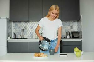 glücklich schön jung blond Mädchen im ein gut Stimmung steht in der Nähe von das Tisch, lächelnd, haben Frühstück, Herstellung Tee im ein groß grau Teetasse, Denken Über das Kommen Tag, auf ein Küche Hintergrund foto